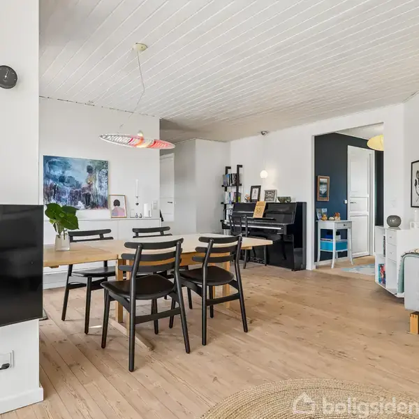 Stue med spisebord, omgivet af sorte stole, og en sofa med grønne planter. I baggrunden ses et klaver og kunst på væggen. Lys fra en stor lampe oplyser rummet.