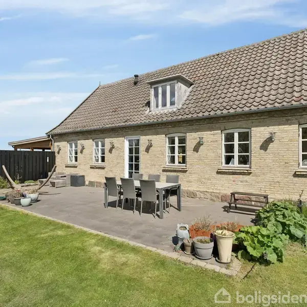 Et murstenshus med skråt tag står i en have. Der er et terrasseområde med spisebord og stole, omgivet af blomsterkrukker. Landskabet er grønt med åbne marker.