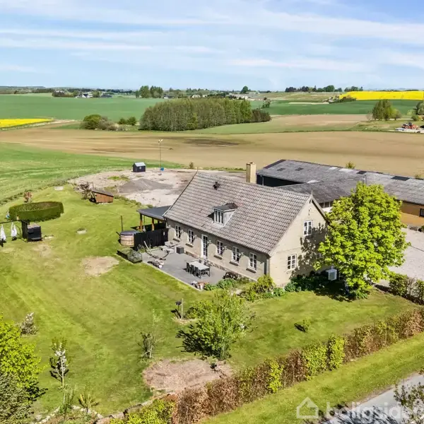 Et stort, gråt hus med have i en landlig omgivelser. Omgivet af marker og træer, grusvej fører til en lade og parkering. Klart, solrigt vejr med spredte skyer.