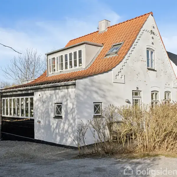 Et hvidt hus med rødt tegltag står i solskin. Huset har flere vinduer, og omgives af grus og buske. Det er i et beboelsesområde.