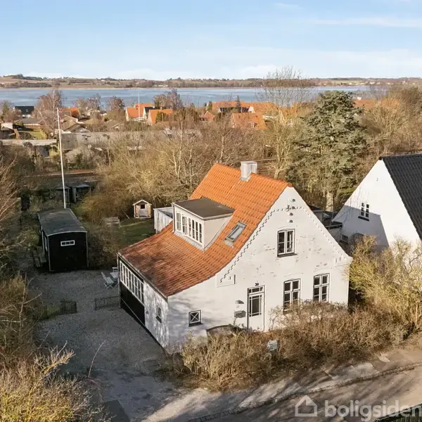 Et hvidt hus med rødt tag står blandt træer, tæt ved en vej. I baggrunden ses en sø og flere huse ligger omkring.