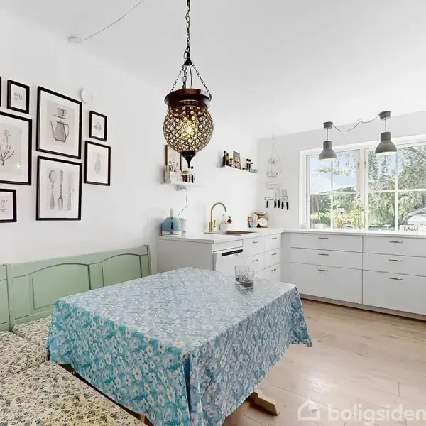 Køkkenbord med blomstret dug foran lysegrøn bænk, under hængende lampe. Billederammevæg til venstre, moderne hvidt køkken med træbordplade og to vinduer, der slipper dagslys ind.