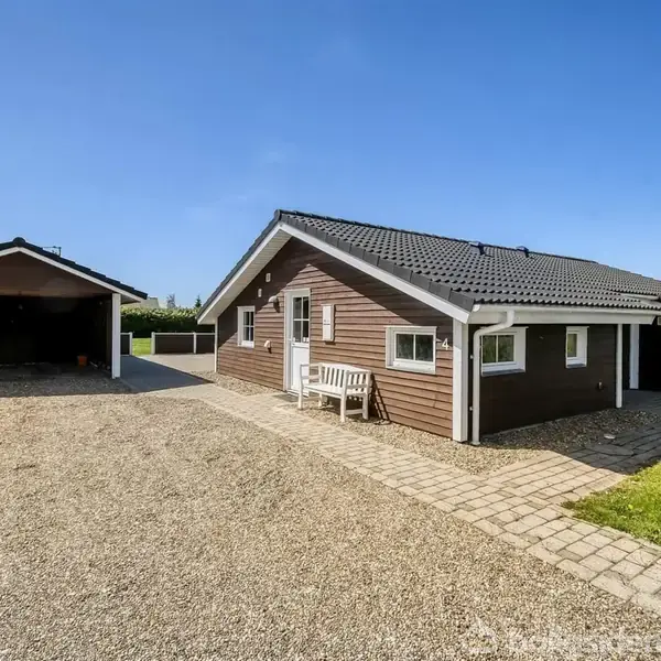 Huset står stille ved siden af en grusvej med en garage til venstre. Sollys skinner på græsplænen, og himlen er klar og blå.