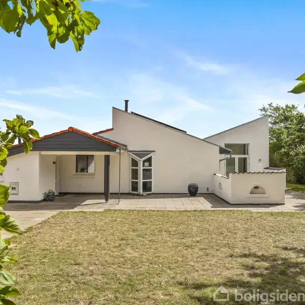 Hvidt hus med moderne design står stille i en græsbeklædt have. En flisebelagt terrasse omgiver bygningen, mens sollys filtrerer gennem omkringstående træer.