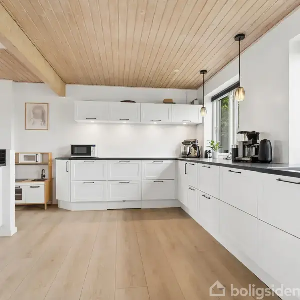 Køkken med hvide skabe og sort bordplade. Kaffeapparat, brødrister og mikrobølgeovn placeret på bordpladen. Store vinduer til venstre, trægulv og træloft skaber lys atmosfære.