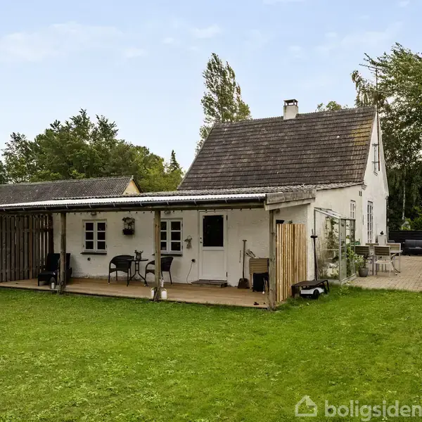 Et hvidt hus med terrasse har havemøbler foran, omringet af en grøn græsplæne og træer. I baggrunden er der en flisebelagt gård med flere møbler.