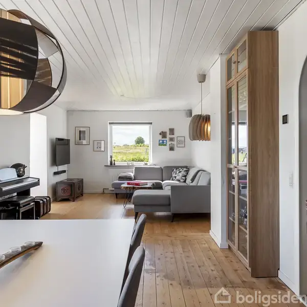 Spisebord med moderne lampe hænger i en stue. Sofa og klaver står nær et vindue med udsigt til natur. Væggene har billeder, og der er trægulve.