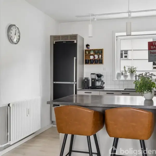 To brune barstole står ved en køkkenø i en moderne køkken. Køleskabet er sort, og der er en vægursklokke til venstre. Et skilt "FOR SALE" ses gennem vinduet.