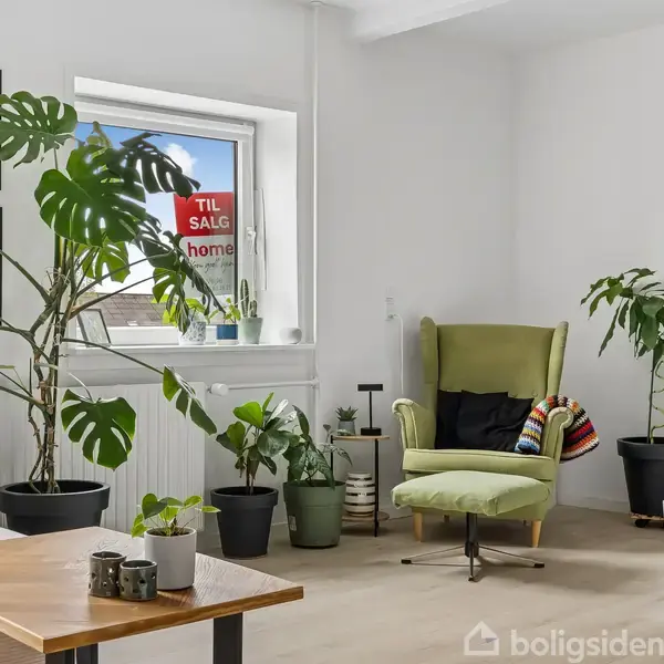 En grøn lænestol står i en stue fyldt med store planter. Et "Til salg" skilt ses i vinduet sammen med farverige vægplakater. En moderne indretning præger rummet.