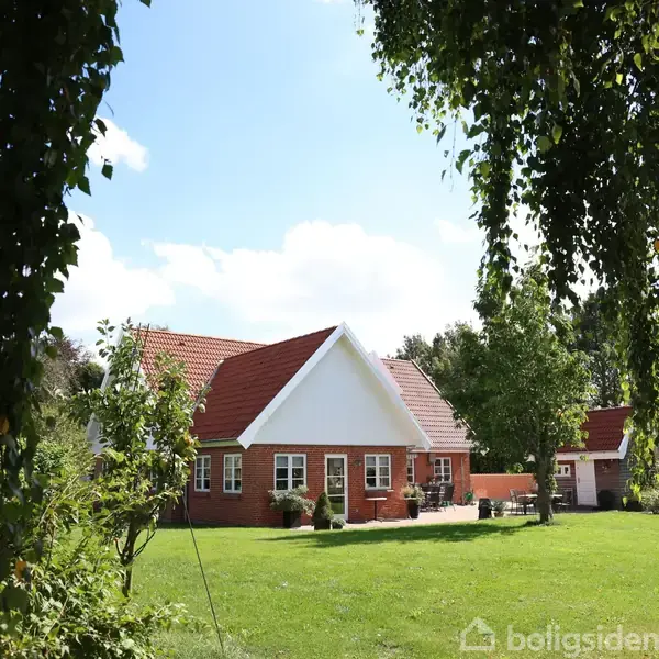 Et rødt murstenshus med hvidt tag og vinduesrammer står omgivet af en grøn græsplæne og træer. Himlen er blå med skyer, og en sti fører til huset.
