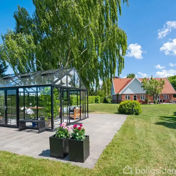 Et glashus står på en terrasse omgivet af en grøn græsplæne med blomsterkrukker foran. I baggrunden ses et hus med rødt tag under en blå himmel.