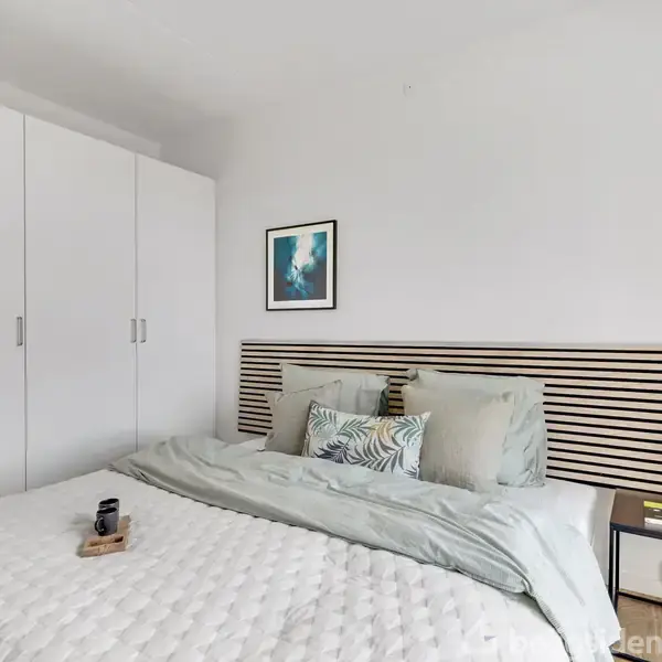 Soveværelse med en stor seng dækket af en hvid dyne og grønne puder. Garderobeskab til venstre, natbord med lampe til højre, og kunstværk på væggen.