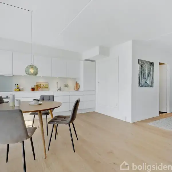 Rundt spisebord med fire stole i et lyst køkken-alrum; moderne indretning med hvidt køkken mod væggen, træ-lys gulv, hængende lampe og dør der åbner til et tilstødende rum.