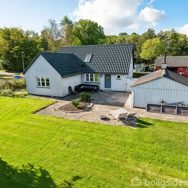 Hvidmalet hus med sort tag står i en stor grøn have. En terrasse med havemøbler ligger ved siden af et skur. Skove omgiver ejendommen i baggrunden.