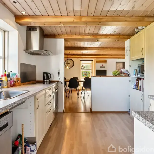 Et køkken med grå bordplader og træloft. Der er en opvaskemaskine, vask, og emhætte. Køleskabet har tegninger på. I baggrunden ses spisebord og stue med ur på væggen.