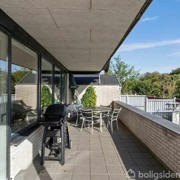 Grill på en flisebelagt altan med bord og stole. Altanen er omgivet af et hvidt murværk og en glasfacade, omkranset af grønne træer i en have.