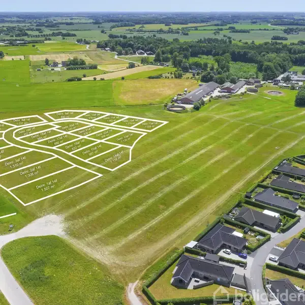 Grønt areal med ensartede marker, set fra luften, omgivet af en lille by med tætliggende boliger og spredte træer under en skyet himmel.