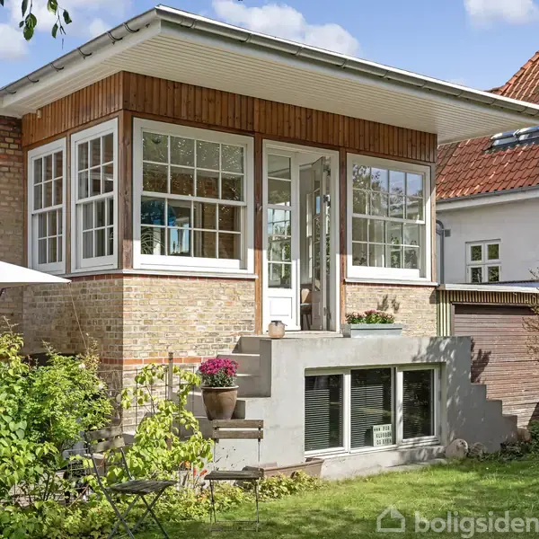 Hus med store vinduer og en terrasse med blomsterkrukker. Underetagen har vinduer, og omgivelserne inkluderer en grøn have med en paraply. Murstensfacade og rødt tegltag.