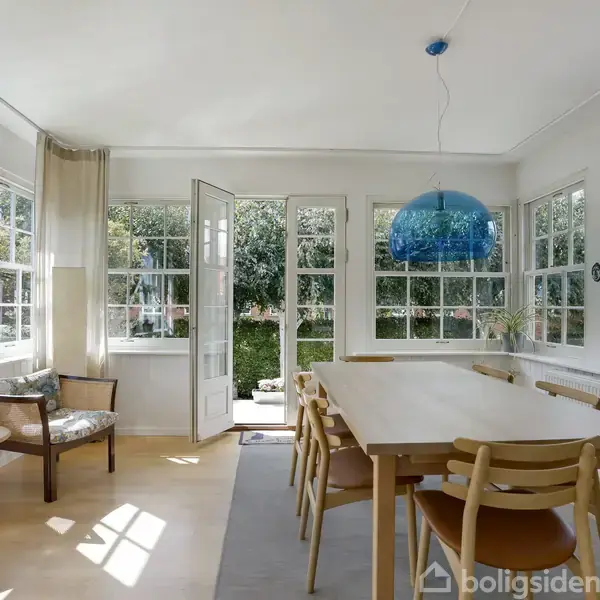 Spisebord af lyst træ med stole står i en lys havestue med mange vinduer. Der hænger en blå lampe over bordet, og døren står åben til en have.