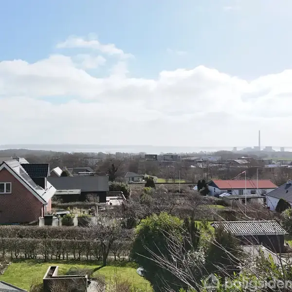 Et hus står stille omgivet af en have med træer og buske i en forstad. I baggrunden ses flere huse og en flad horisont under en let overskyet himmel.
