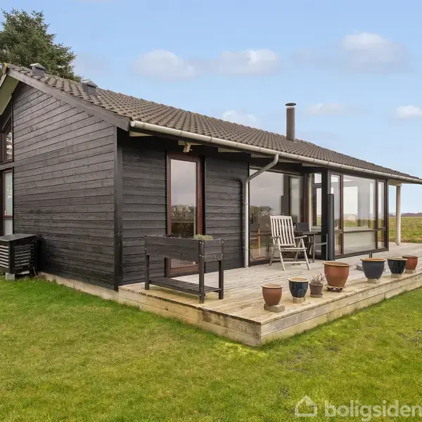 Et sortmalet træhus står på en græsplæne. Huset har en terrasse med flere blomsterpotter og en stol, omgivet af åbent land.
