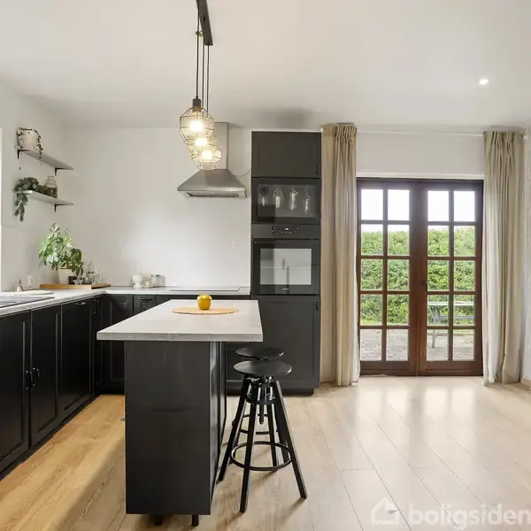 Køkken med sort køkkenø og stol, har hængende lamper. Store vinduer og døre åbner mod en have. Træskab står til højre. Planter tilføjer grønt.