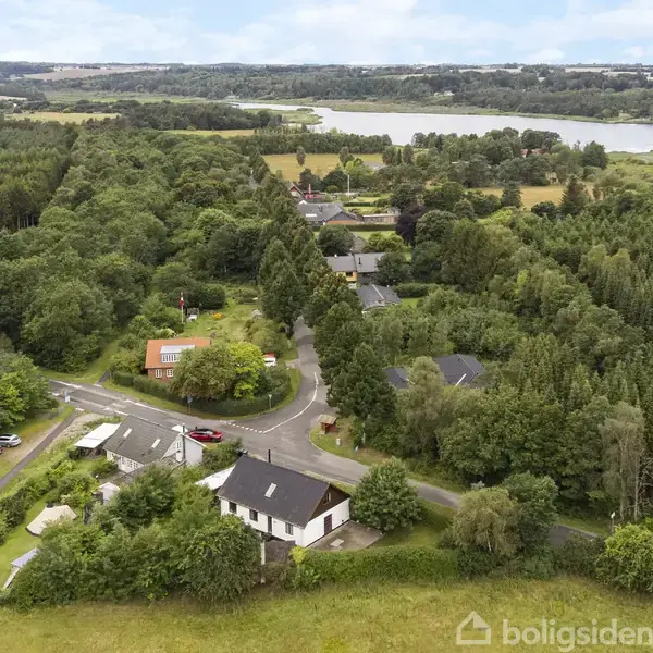 Et hus står blandt grønne træer nær en vej i et skovområde. Området er frodigt med skove og marker, og en sø er synlig i det fjerne.