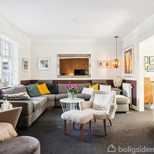 En stue med en stor grå sofa dekoreret med farverige puder. Der er et centralt bord med blomster, omgivet af lænestole. Lys træ-konsol ved siden af lamper og billeder.