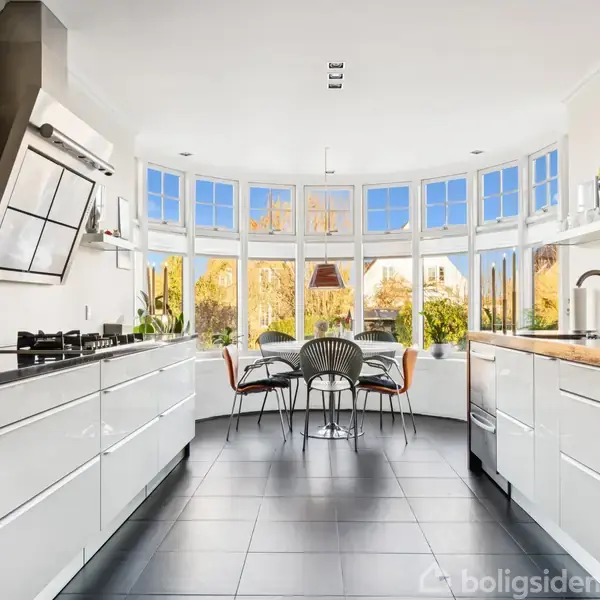 Et moderne køkken med hvide skabe og sorte fliser. En rund spisebordsgruppe står ved store buevinduer, der giver udsigt til en have med efterårsfarver.