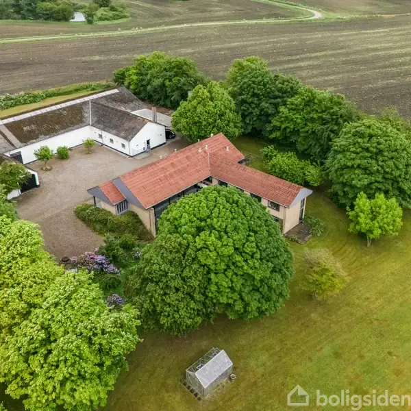 Et landligt hus med rødbrunt tag er omgivet af tæt skov og marker. Et mindre udhus og gårdsplads er synlige. Grønne træer omkranser bygningerne, der ligger i et landbrugsområde.
