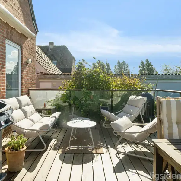Udendørs terrasse har komfortable stole omkring et rundt bord. En gasgrill står til venstre. Plantepotter og træhegn omkranser området, mens en klar himmel ses i baggrunden.