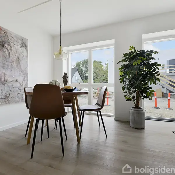 Et spisebord med stole står i et lyst rum. Der hænger et kunstværk på væggen, og store vinduer viser en moderne bygning udenfor. En plante står ved døren.