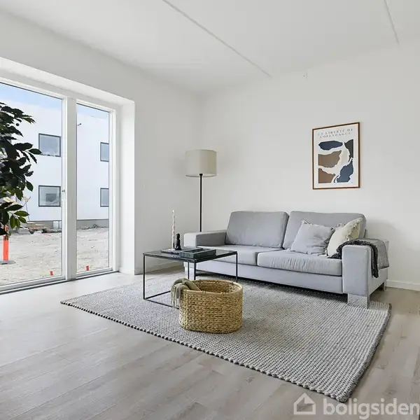 Sofa står roligt i et lyst og minimalistisk stueområde med trægulv. Tæt ved vinduet står en stor plante og en gulvlampe. Et billede hænger på væggen.