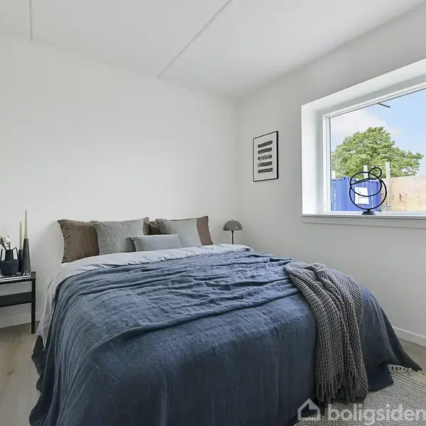 En seng dækket med blå sengetøj står stille i et lyst soveværelse. Ved siden af er et natbord med en plante og lampe. Et vindue viser udsigt til træer udenfor.