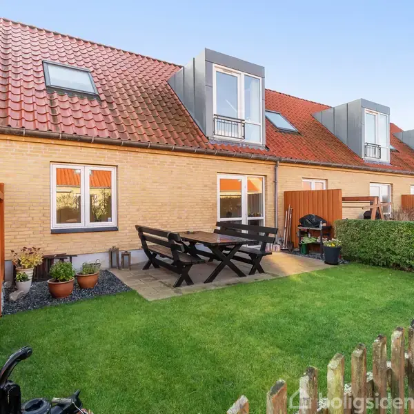 En terrasse med havemøbler står foran et rækkehus med røde tage. Der er en velholdt græsplæne, blomsterpotter, og en lav træhegn omkring haven.