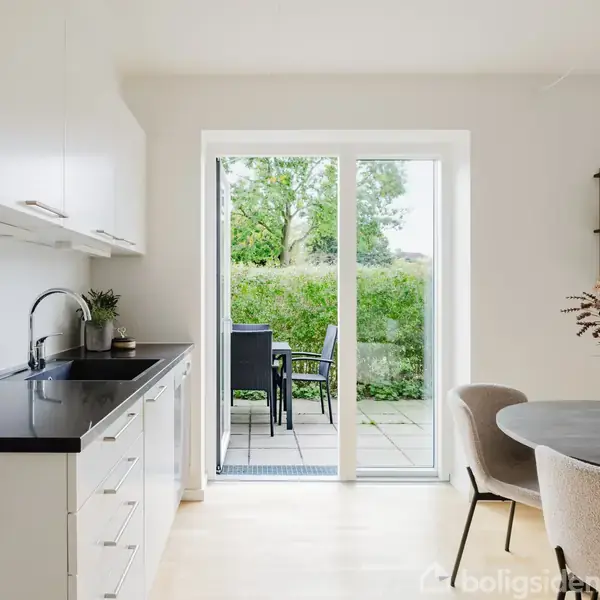 Et moderne køkken med hvidt interiør og sort bordplade, vender mod en glasdør, som åbner til en terrasse med grøn udsigt. Rund spisebord med blomsterdekoration, stol omkring.