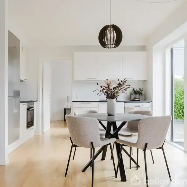 Et spisebord med fire polstrede stole står i et lyst, moderne køkken. En vase med tørrede blomster er placeret på bordet. Store vinduer giver udsigt til en grøn have.