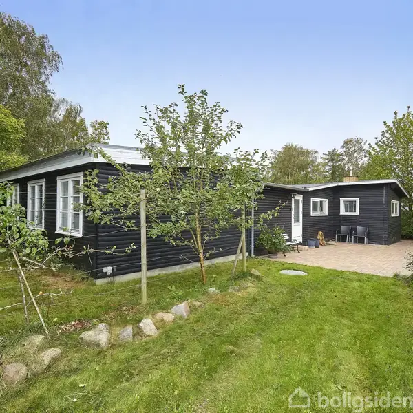 Et sortmalet hus ligger på en grøn græsplæne, omgivet af træer. En træterrasse med svømmebassin er synlig ved siden af huset, omgivet af en rolig, naturlig have.