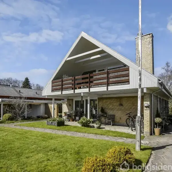 Et moderne hus med spidst tag og altan står i grøn have med flisebelagt sti. En garage ses til venstre, mens cykler står under taget. Skyfri himmel i baggrunden.