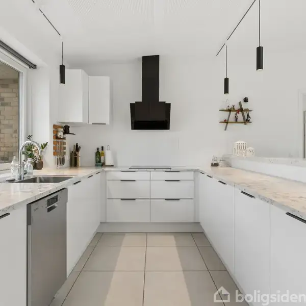 Et moderne, lyst køkken med hvide skabe og marmorflisebordplader. En sort emhætte hænger over komfuret. Store vinduer giver naturligt lys. Køkkenet har indbyggede apparater og minimalistisk indretning.