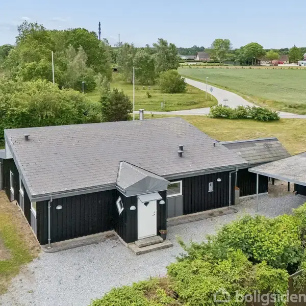 Sort hus med fladt tag står stille på en grusbelagt oplads. Ved siden af er en carport. I baggrunden ses åbne marker og træer, nær en snoet vej.