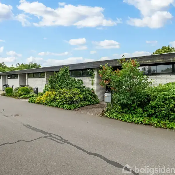 Rækkehus bygninger langs en asfalteret vej med tæt beplantning. Skyer på blå himmel danner baggrund.
