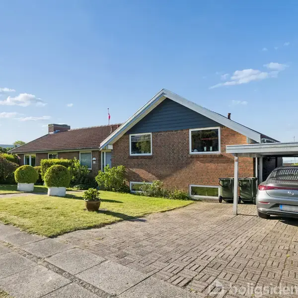 En villa med murstensfacade og græsplæne, står under en blå himmel. Foran huset er en carport med en parkeret bil. Græsplænen er velplejet med trimmet hæk.