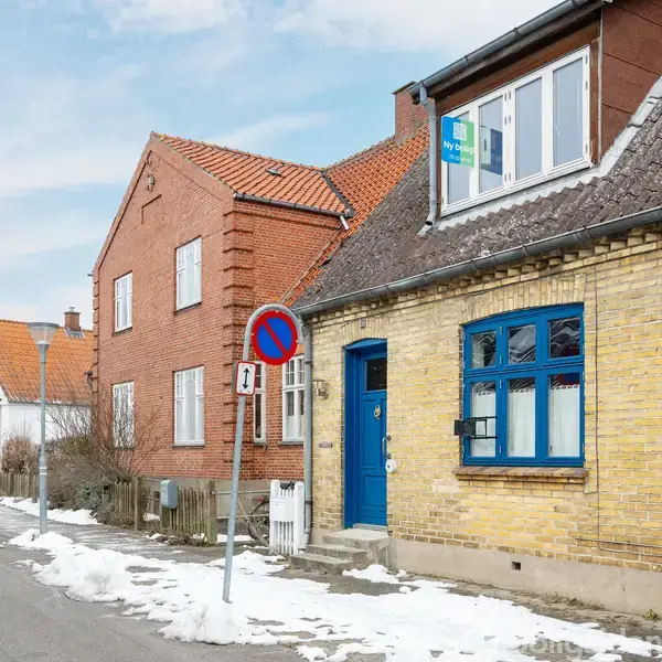 En beige murstenshus med blå dør og vinduer står ved en sneklædt vej. Et "ingen parkering" skilt er synligt. Andre huse med røde tegltage ses i baggrunden.