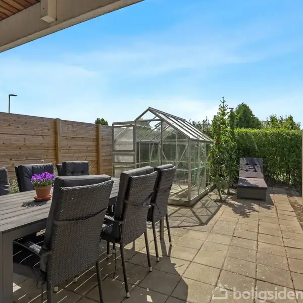 En terrasse med spisebord og seks stole, der står ved siden af en grill. Et lille drivhus ses i baggrunden omgivet af en træhegn og grønne buske.