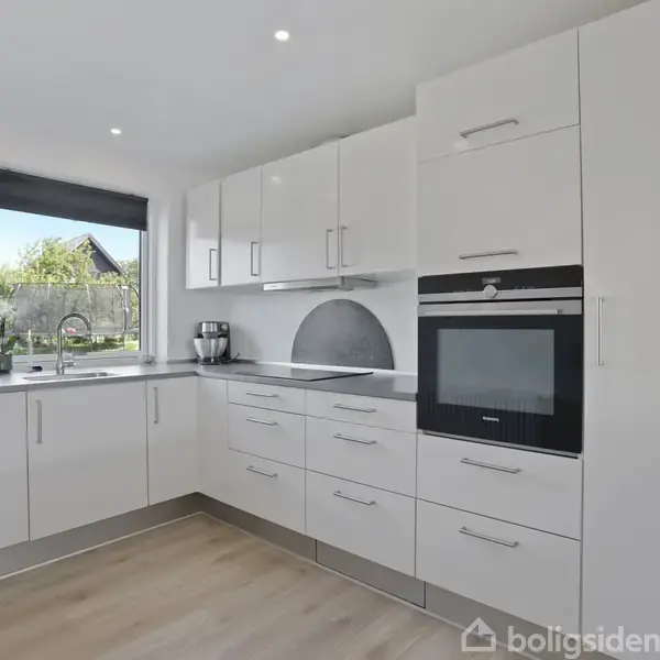 Et moderne køkken med hvide skabe og en indbygget ovn i højre side. En kaffemaskine står på bordpladen ved vinduet, som giver udsigt til en have.