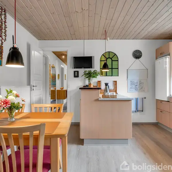 Spisebord med blomsterbuket står i lyst køkken/alrum. Træloft og lyse trægulve præger rummet. Køkkenet har moderne apparater og dekorationer på væggene, som indeholder billeder og et ur.