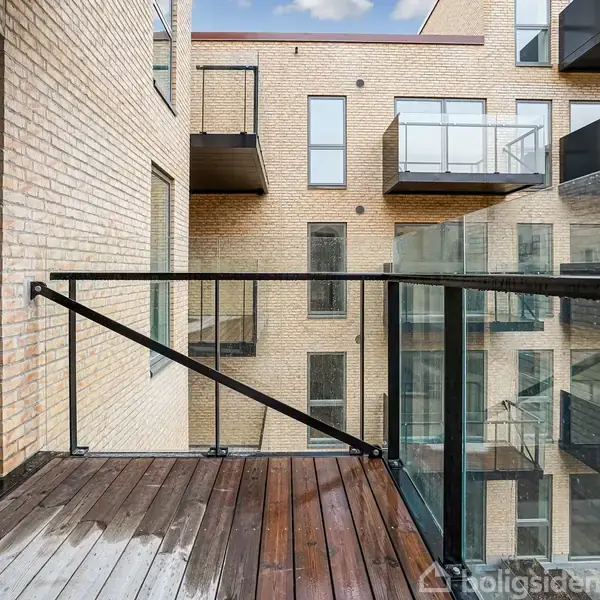 En balkon med trægulv og glasrækværk vender mod en moderne lejlighedsbygning med store vinduer. Bygningen er af lyse mursten, og flere balkoner ses i baggrunden.