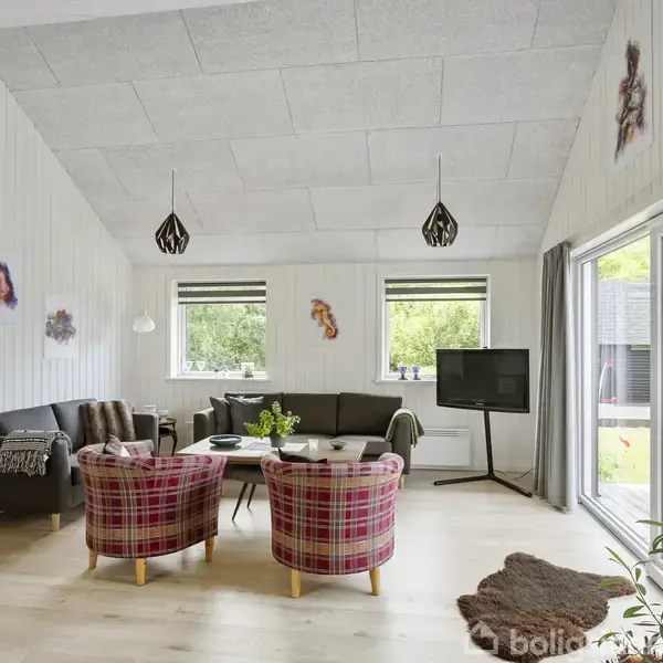 Stue med to ternede stole og en sort sofa rundt om et sofabord. Et fjernsyn står ved siden af vinduerne, som viser en have udenfor.