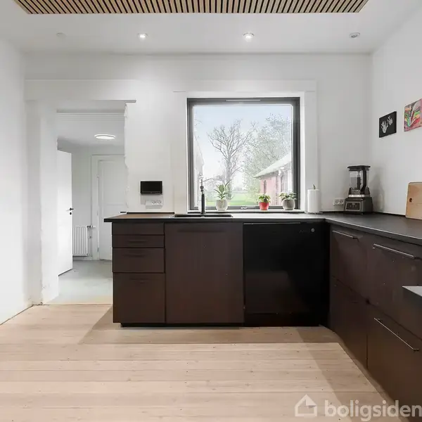 Køkkenet har mørke skabe og en sort bordplade. Der står en blender og skærebræt på disken. Et vindue åbner sig til en have. Væggene er hvide.